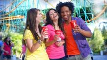 Three teenagers walking in the park sipping a soda with roller coaster in background